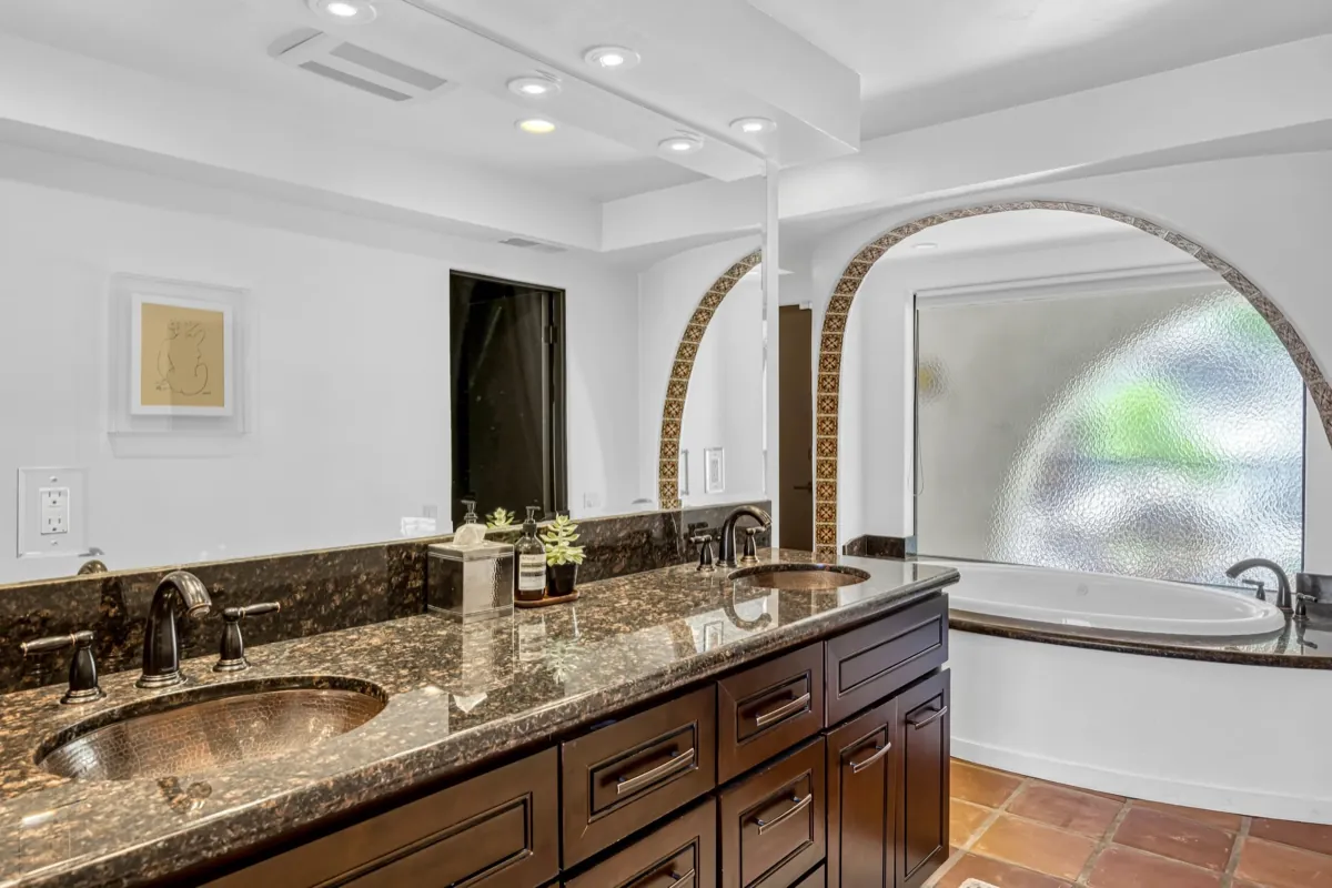 Double vanity in dark granite with an arched brick window above the soaking tub.