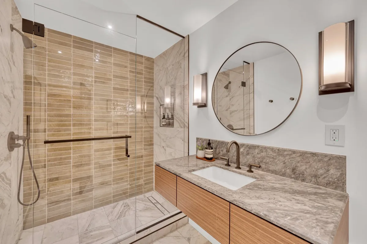 Marble bathroom with a glass shower, round mirror and a wide stone vanity.