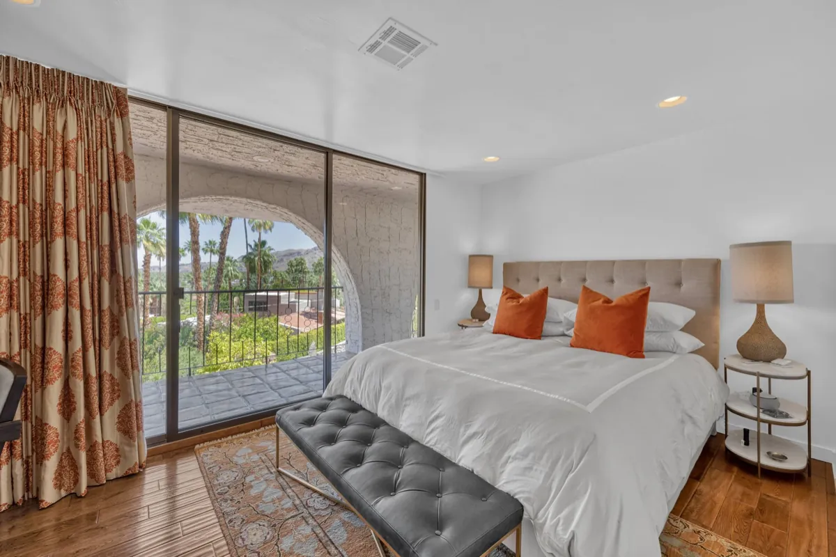 Guest bedroom with orange accent pillows and a tufted bench, opening to an arched window and palms.