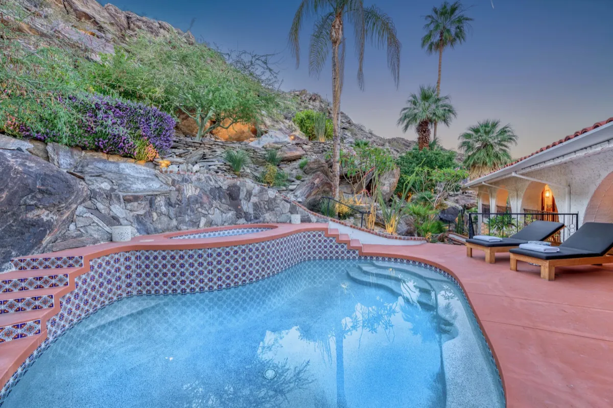 The pool at twilight below the boulder hillside, palms and the lit house behind it.