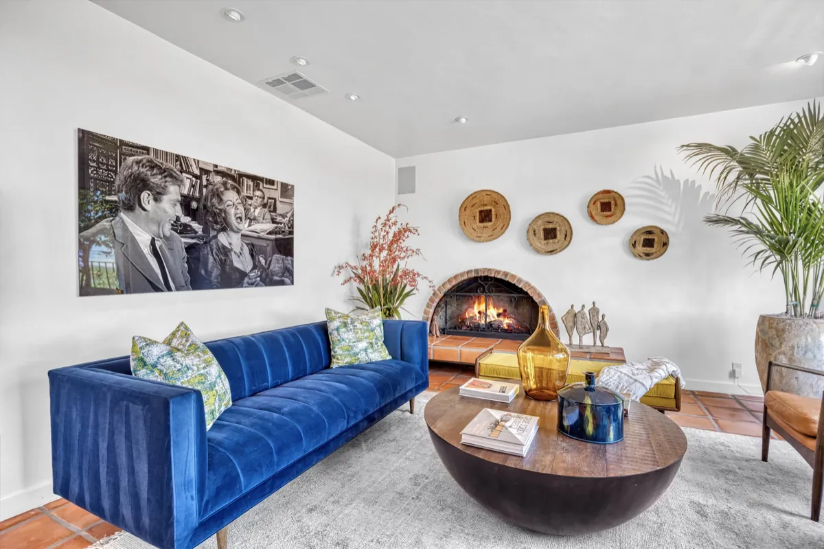Living area with a blue velvet sofa facing a lit corner fireplace and framed artwork.
