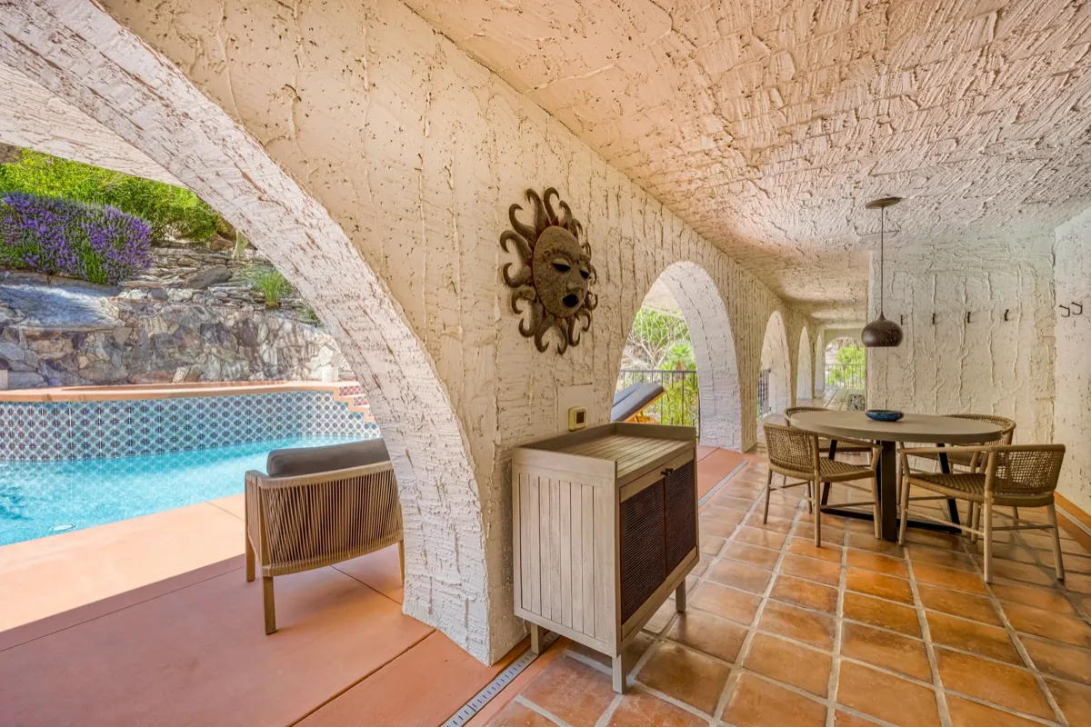 Shaded arched loggia beside the pool with a lounge chair and a wood cabinet.